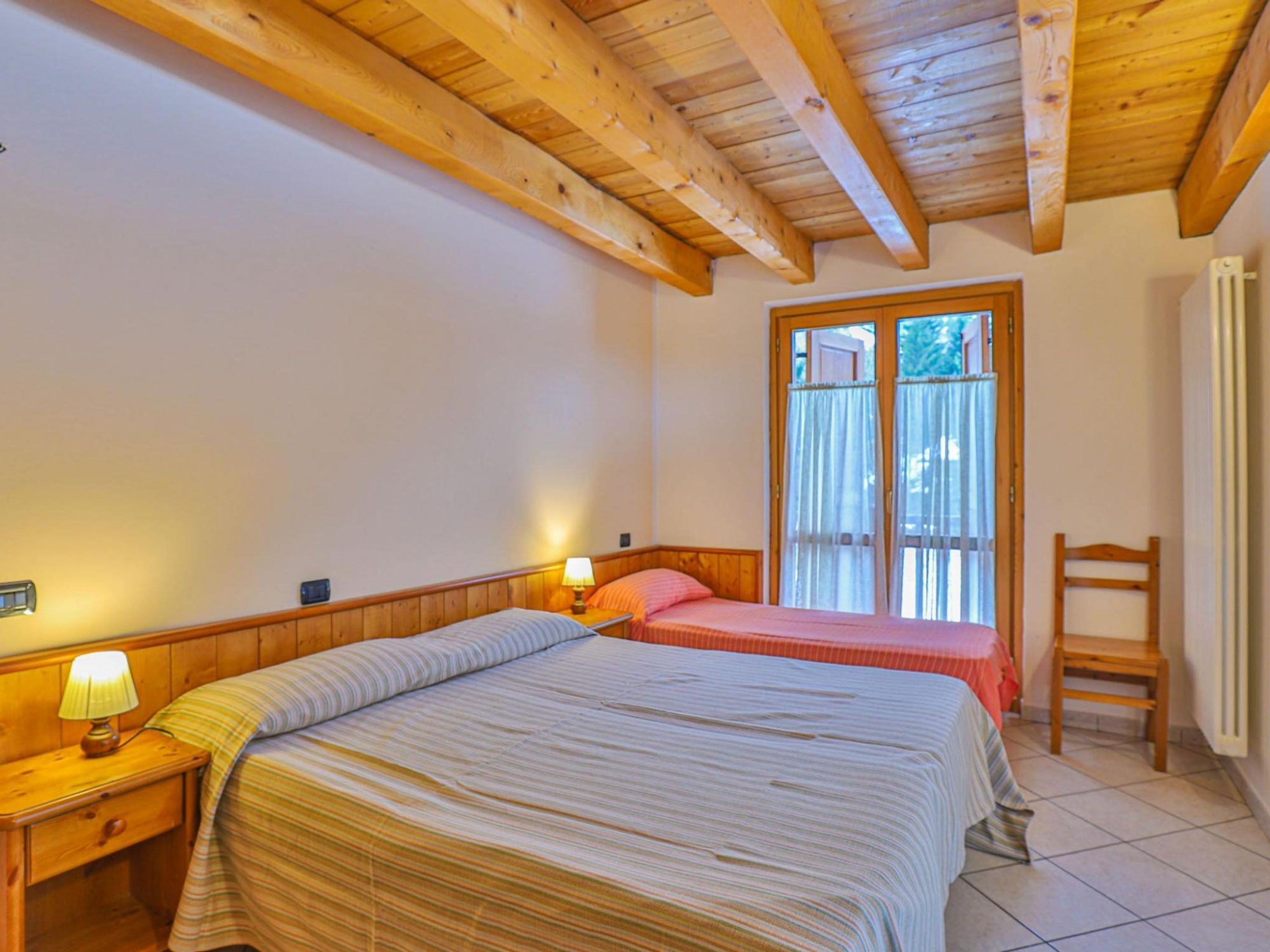 A bedroom with two beds, wooden headboards, a nightstand with a lamp, and a window with curtains.