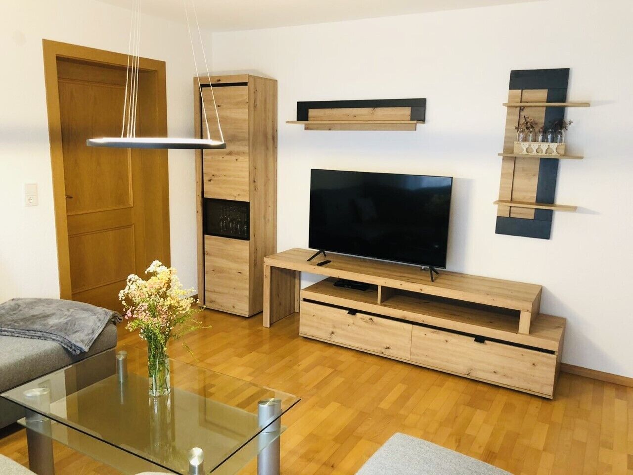 Un soggiorno moderno con un mobile TV in legno, una TV a schermo piatto e un tavolino da caffè in vetro con un vaso di fiori.