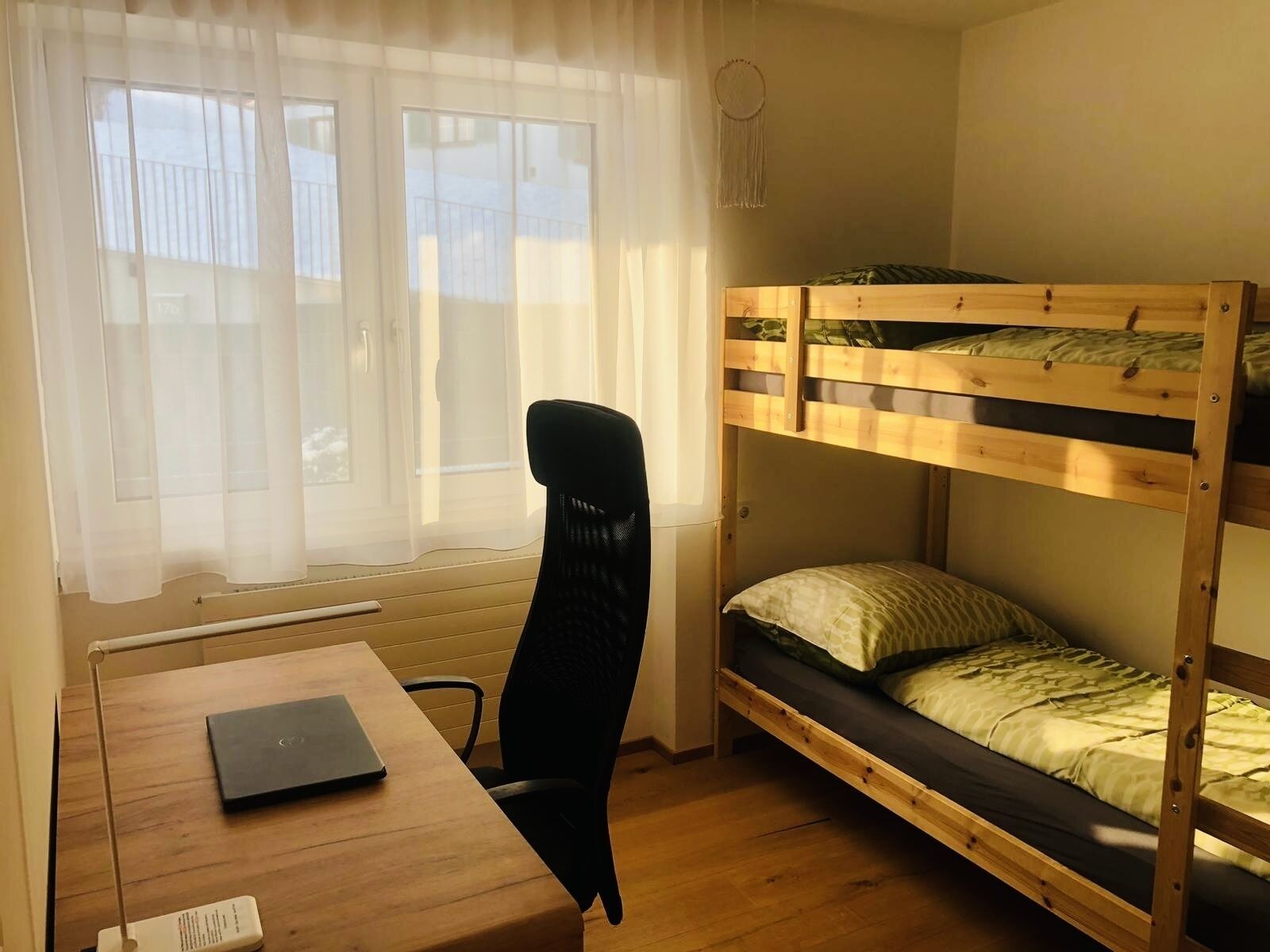 Un llit amb literes, una taula amb cadira, un ordinador portàtil i una finestra amb cortines fines.