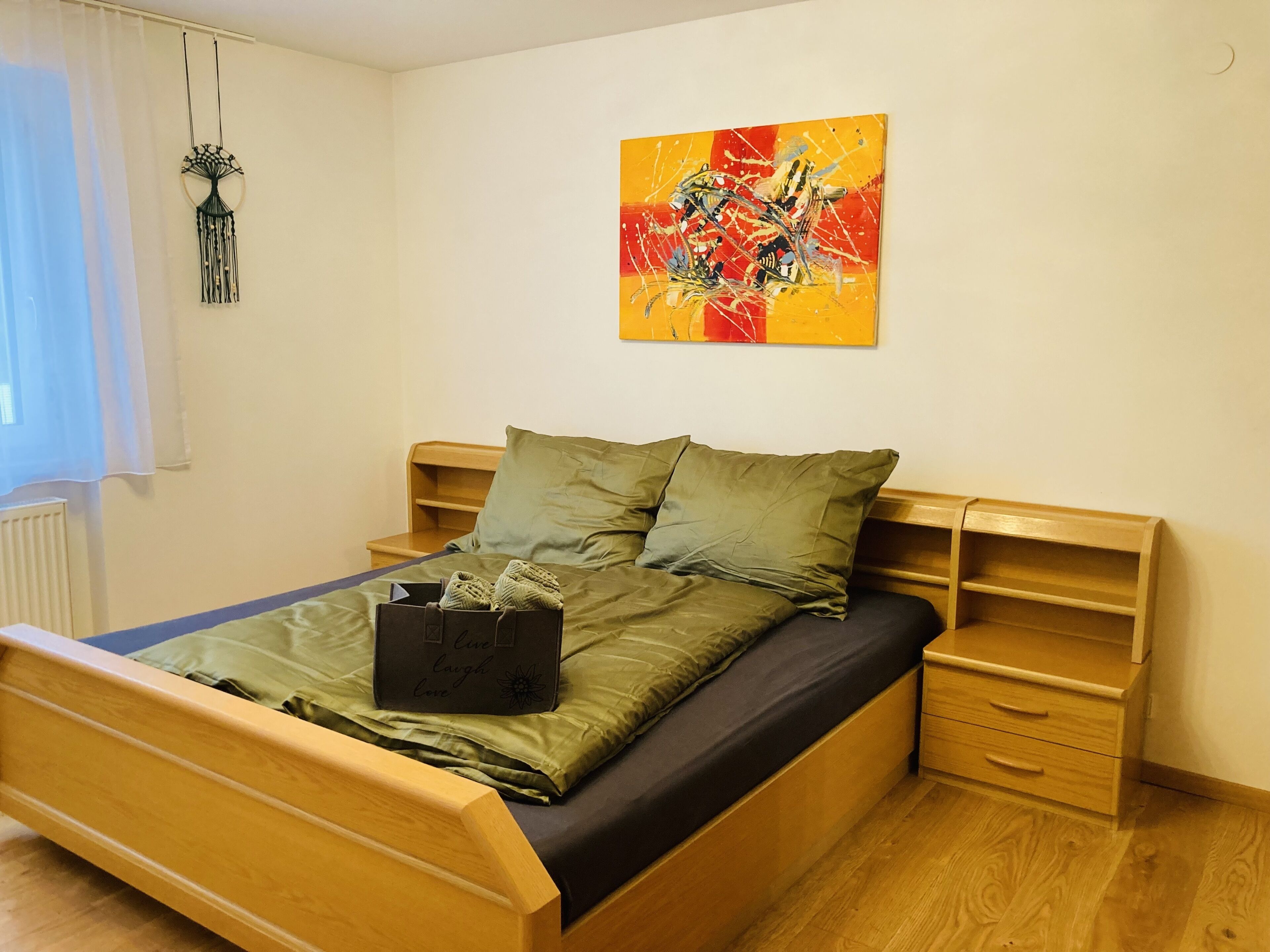 Un lit avec une couverture et des oreillers verts, une tête de lit en bois, une table de chevet avec des tiroirs et un tableau abstrait accroché au mur.