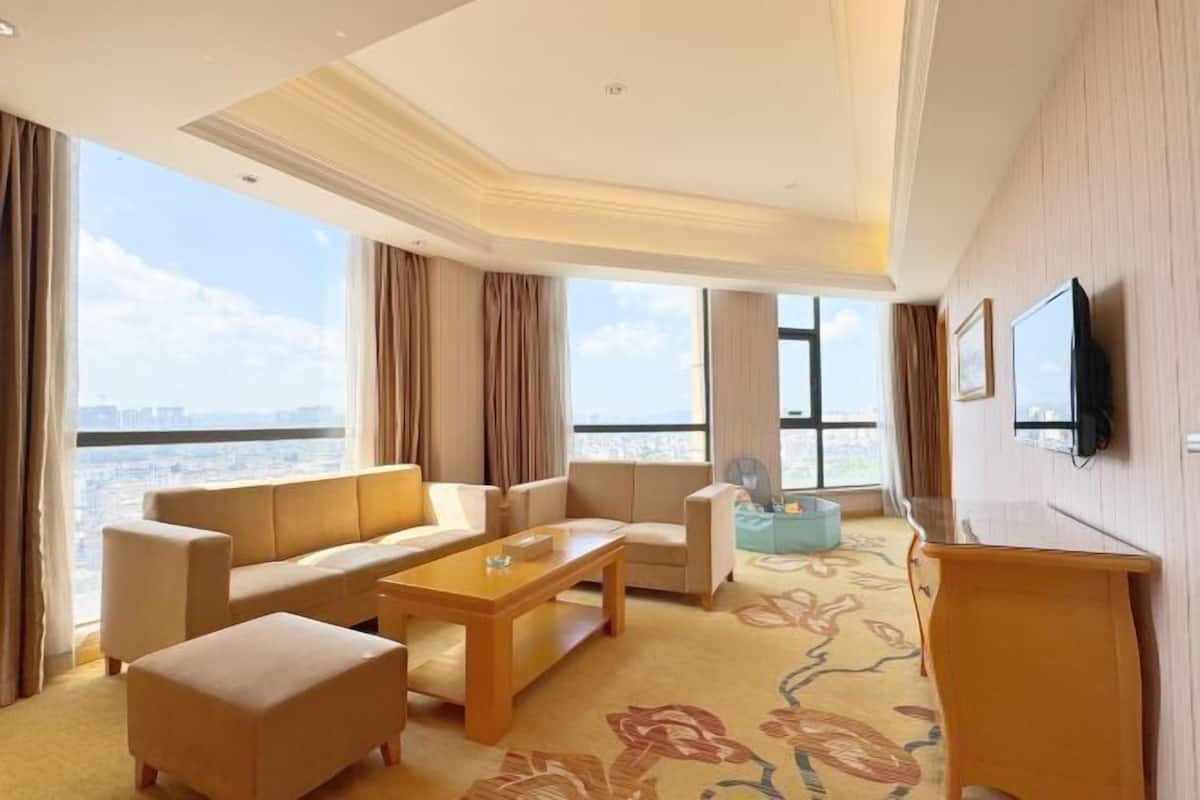 A hotel room with a sofa, armchairs, a coffee table, a TV, and a view of the city.