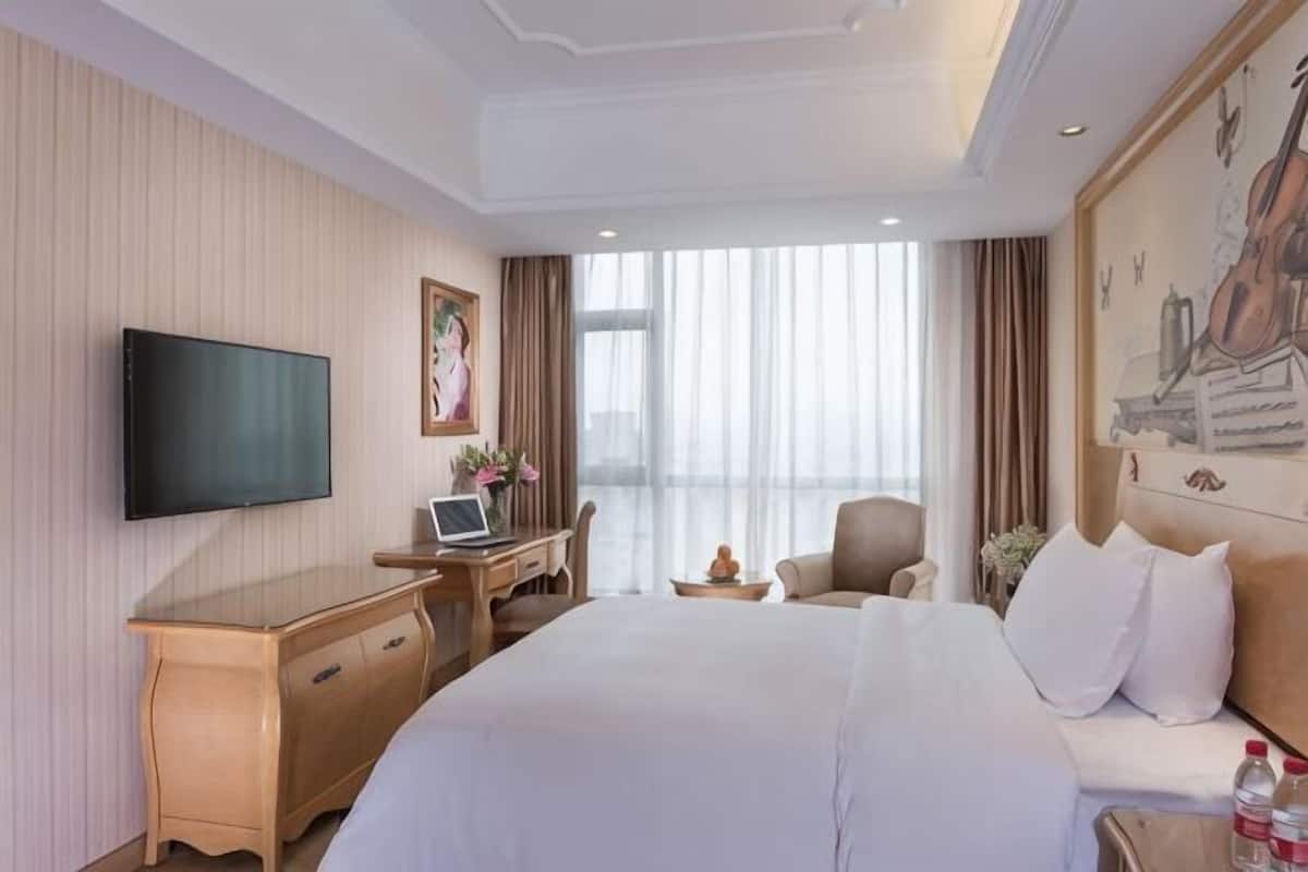 A hotel room with a large bed, a desk with a laptop, a TV, and a painting on the wall.