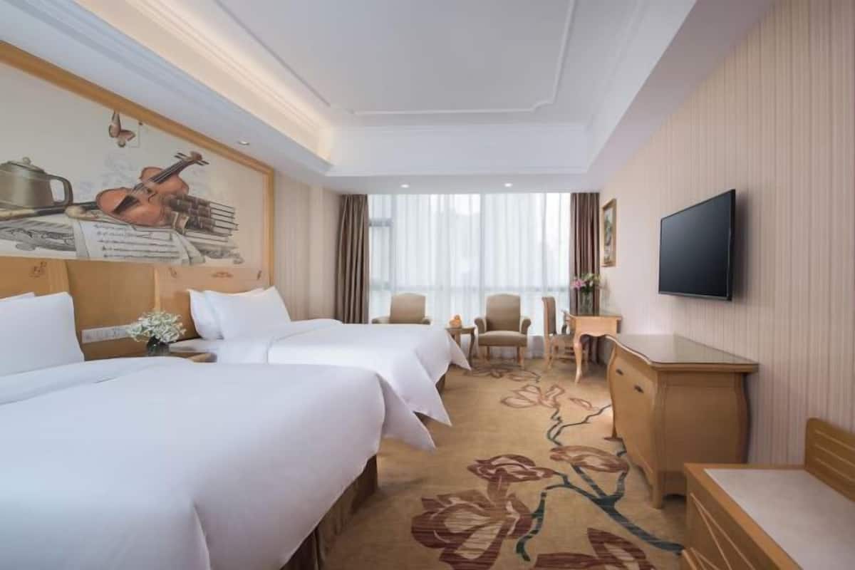 A hotel room with two beds, a flat-screen TV, a desk, and a large window with curtains.