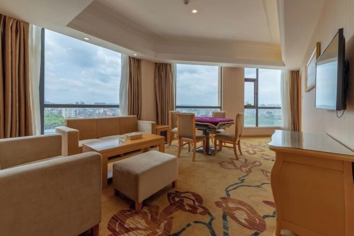 A hotel room with a large window, a flat-screen TV, a dining table, and a sofa.