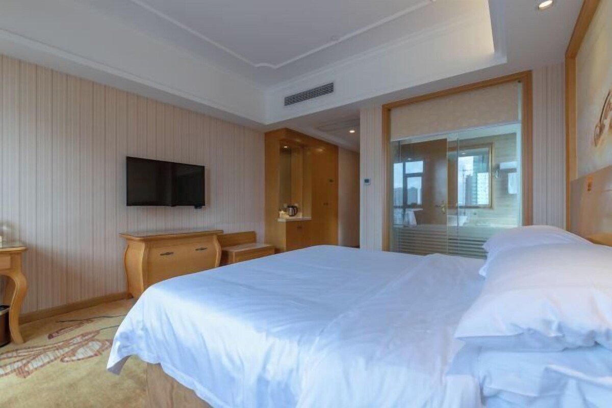 A hotel room with a large bed, a flat-screen TV mounted on the wall, a wooden dresser, and a glass door leading to a balcony.