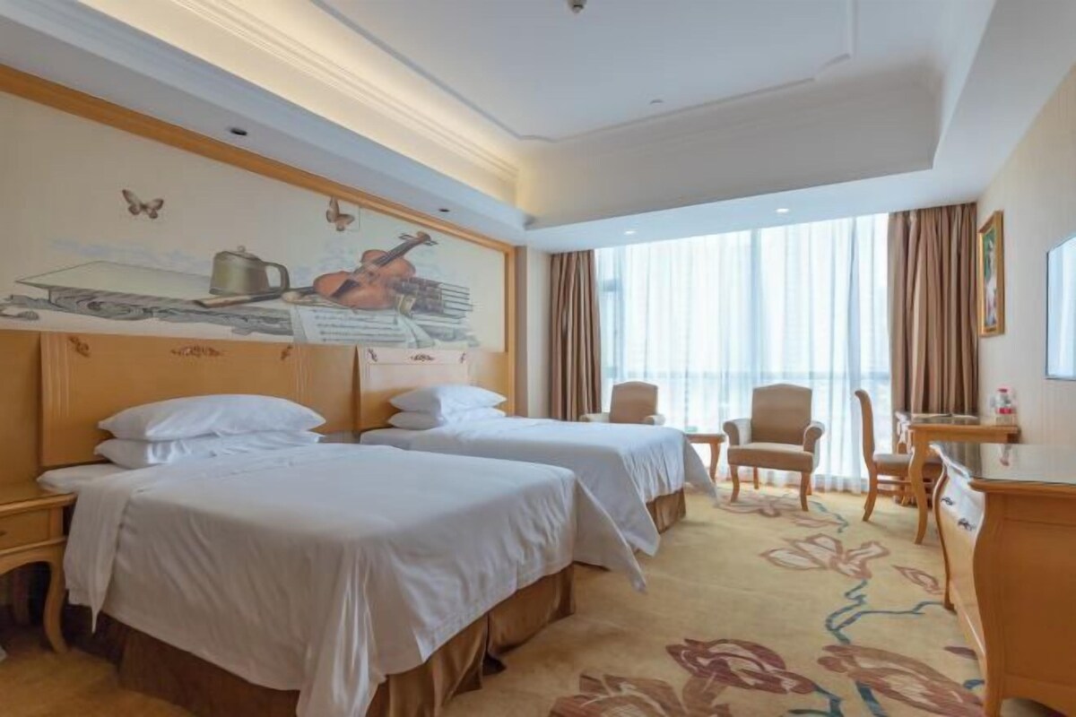 A hotel room with two beds, a desk, a chair, and a large window with curtains.