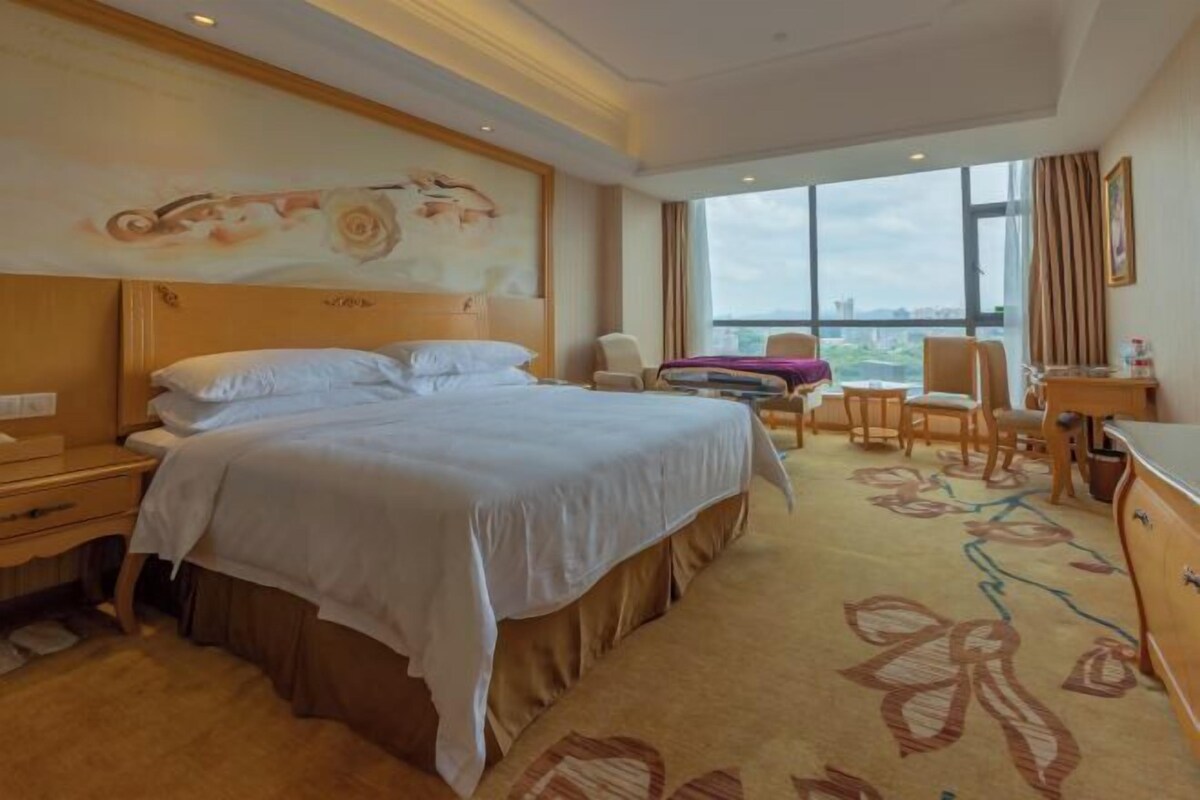 A hotel room with a large bed, a desk, a chair, and a view of the city.
