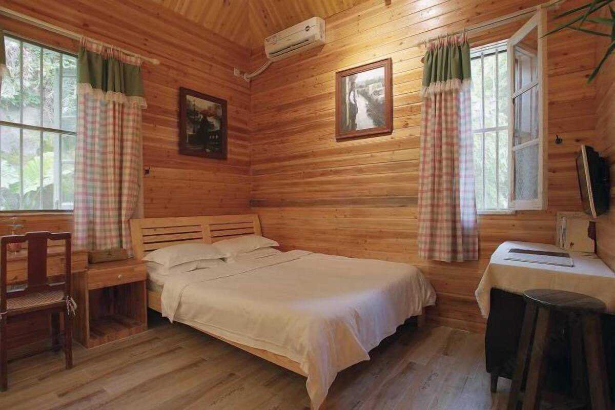 Una habitación de cabaña de madera con una cama, un escritorio, una silla y dos ventanas con cortinas.