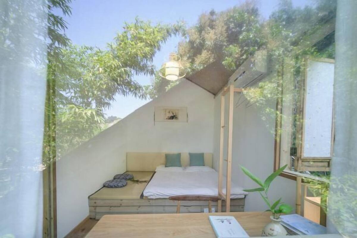Una cabaña pequeña y de una sola habitación con una cama, una mesita y una ventana con vistas a la vegetación.