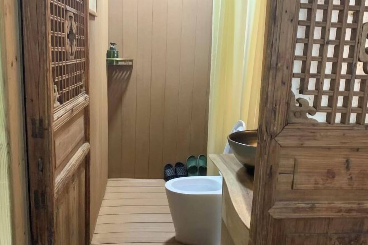 Un estrecho pasillo de madera con una puerta de madera a la izquierda, un inodoro blanco a la derecha y un estante de madera con un cuenco y pantuflas.