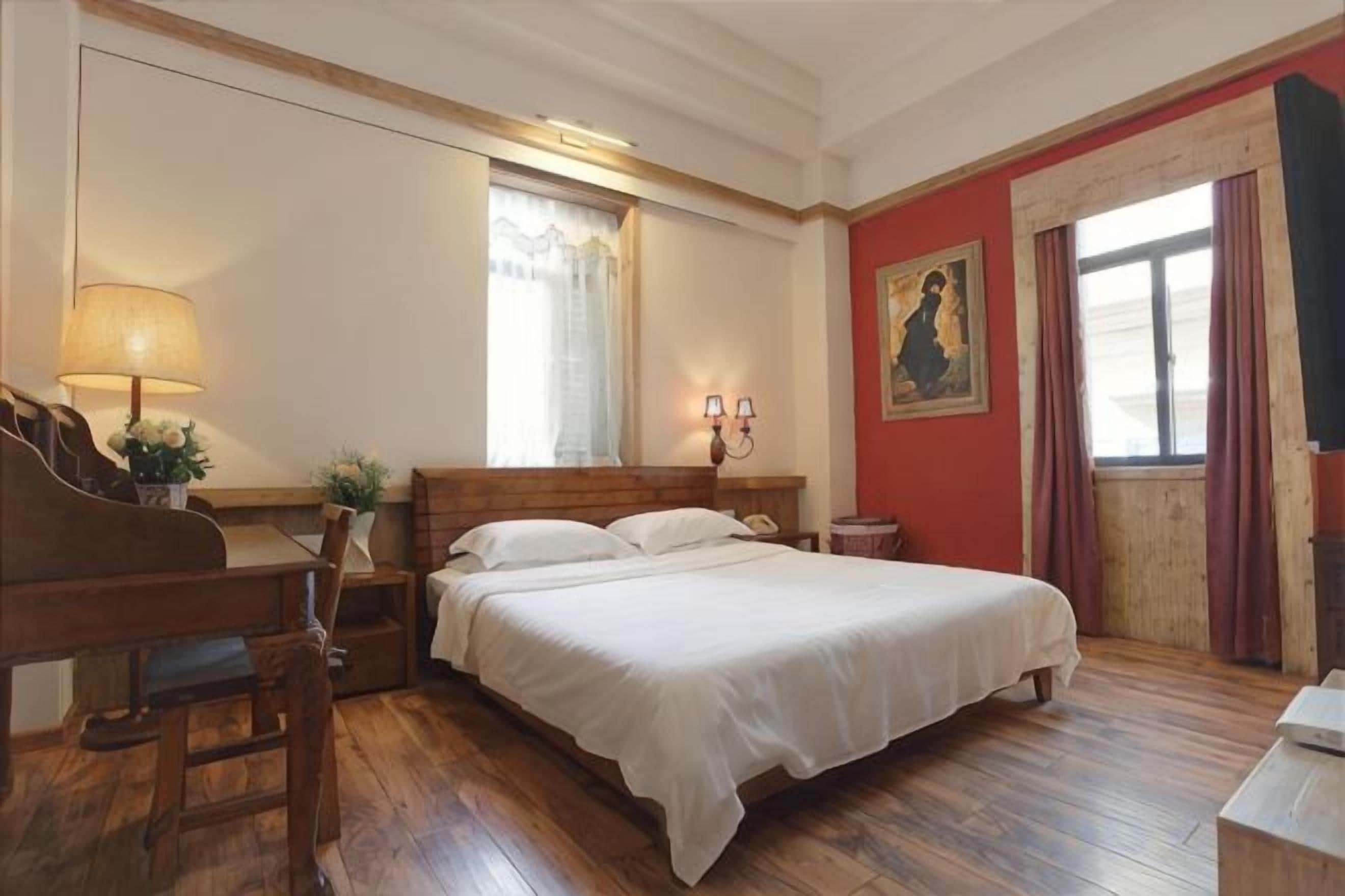 A bedroom with a wooden bed, a desk with a lamp, a window with curtains, and a painting on the wall.