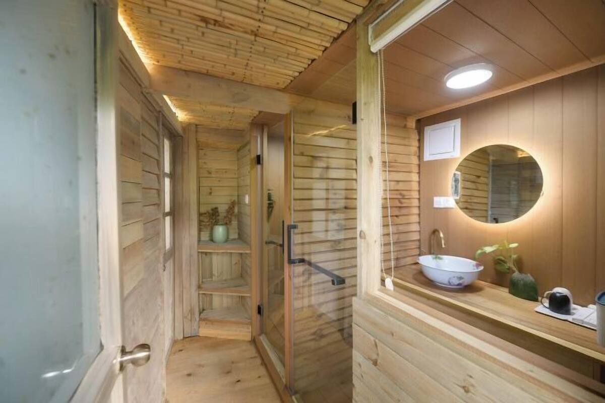 Un sauna de madera con puerta de vidrio, un espejo redondo y un lavabo con un recipiente.