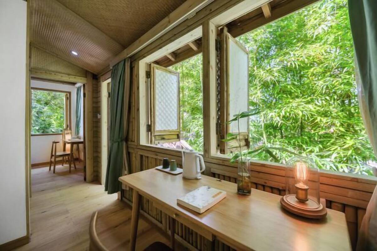 Interior de madera con una mesa, sillas, una ventana que da a vistas de follaje y una lámpara de mesa.