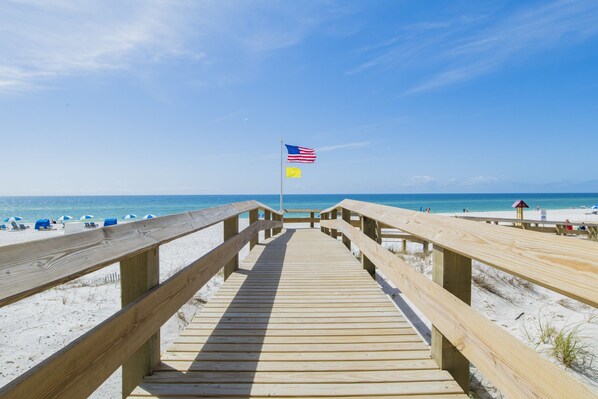A wooden boardwalk leading to a beach with a flag, beach chairs, and umbrellas.