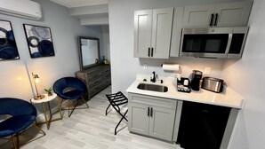 A modern kitchen with grey cabinets, a stainless steel sink, a microwave, and a coffee maker. There are two blue chairs and a small table with a lamp.