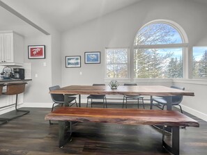 Un comedor con una mesa y bancos de madera, fotografías enmarcadas en la pared y vistas a árboles a través de amplios ventanales.