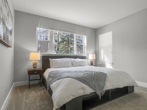 Un dormitorio con una cama, dos lámparas de noche, un ventanal con vistas a árboles y un cuadro en la pared.