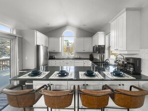 A modern kitchen with white cabinets, a large island with bar stools, and a view of the outdoors through a large window.