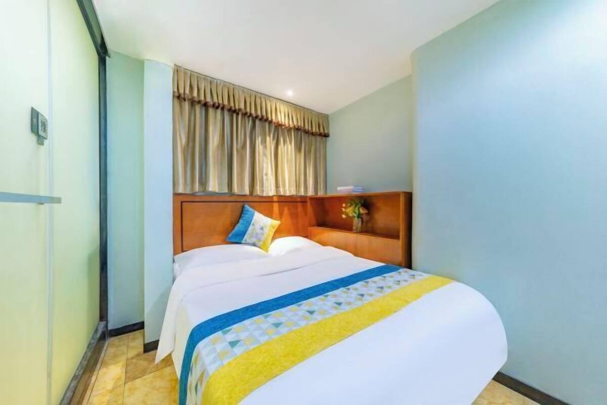 Habitación de hotel con cama, cabecera de madera, cortina y mesita de noche con un jarrón de flores.