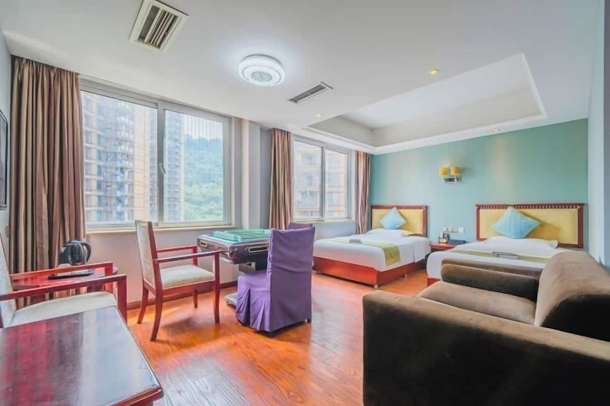 Habitación de hotel con dos camas, un escritorio, una silla y ventanas amplias con vista a la ciudad.