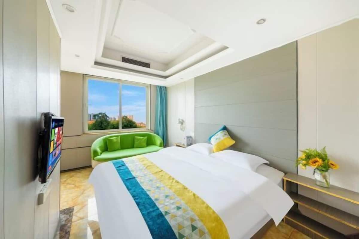 Habitación de hotel con una cama grande, un sillón verde, televisión y ventana con vistas a la ciudad.