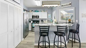 A modern kitchen with white cabinets, a central island with bar stools, stainless steel appliances, and decorative items. - Pool Just Steps Away | Easy Walk to Private Beach & Gulf Coast Fun (Destin)