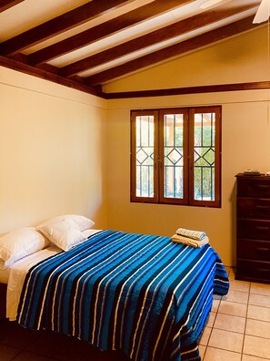 Relax in this serene room with a comfortable bed, a dresser, and a window offering a view of lush greenery.