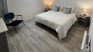 Un dormitorio moderno con tragaluz, una cama con ropa de cama blanca, dos mesitas de noche con lámparas, una silla azul y un escritorio blanco.
