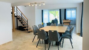 Un comedor con una mesa rectangular y ocho sillas, una escalera y ventanas amplias con cortinas.