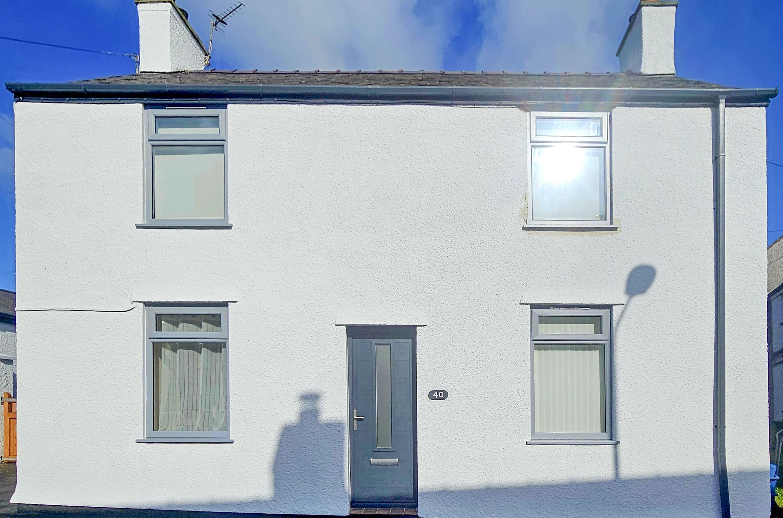 Une maison blanche à deux étages, avec une porte grise, deux fenêtres au rez-de-chaussée et une cheminée.