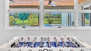 Una habitación con mesa de fútbol, ventanas amplias con persianas y ventilador de techo.