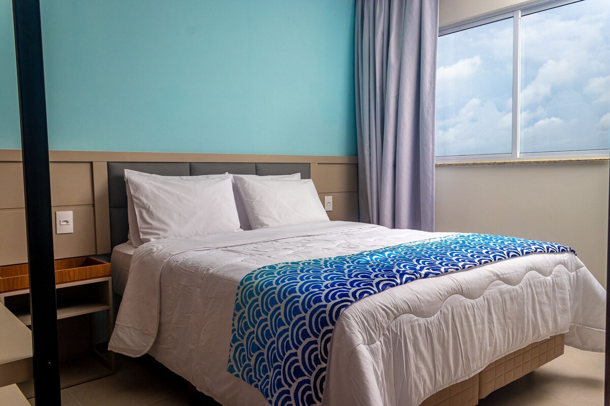 A neatly made bed with a patterned bedspread, a bedside table, and a window with curtains.