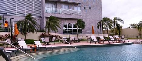 A hotel with a swimming pool, lounge chairs, and palm trees.
