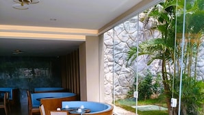 A modern dining area with round tables and blue cushioned seating, large windows with a view of greenery, and a stone-textured wall. - Aqualand Park & Resort Oficial (Salinópolis)