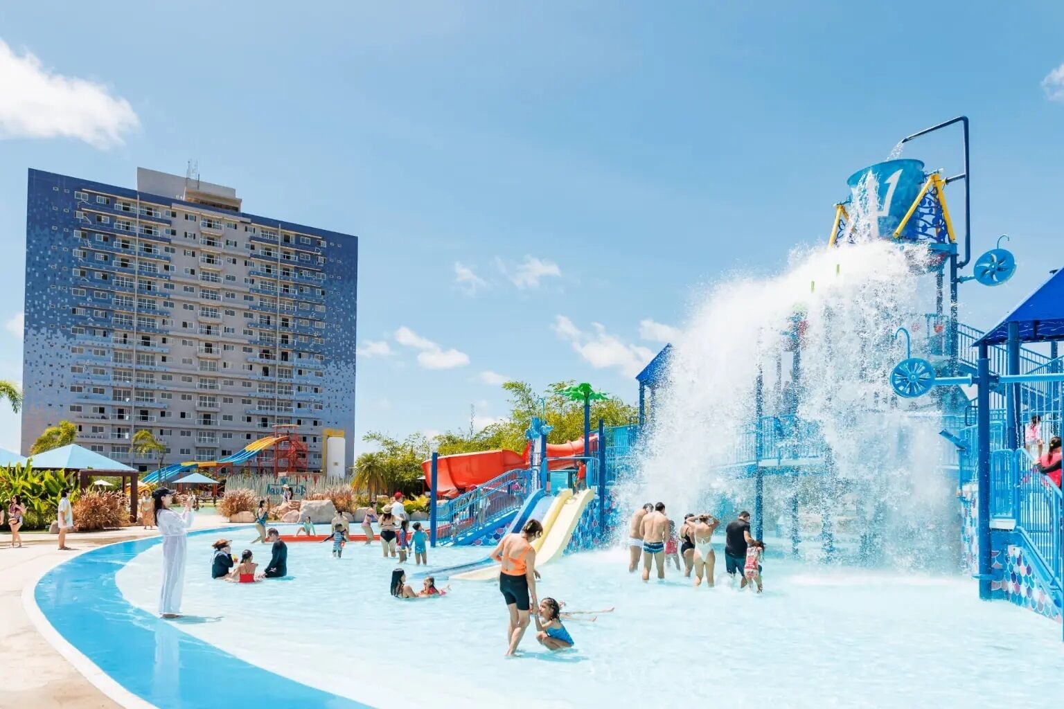 Un parque acuático con un edificio grande al fondo, un tobogán de agua y gente disfrutando de las instalaciones.