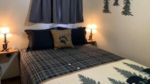 Een slaapkamer met een bed, twee bedlampjes, een raam met gordijnen, en decoratie aan de muur, waaronder silhouetten van bomen en een kussen met een pootafdruk.