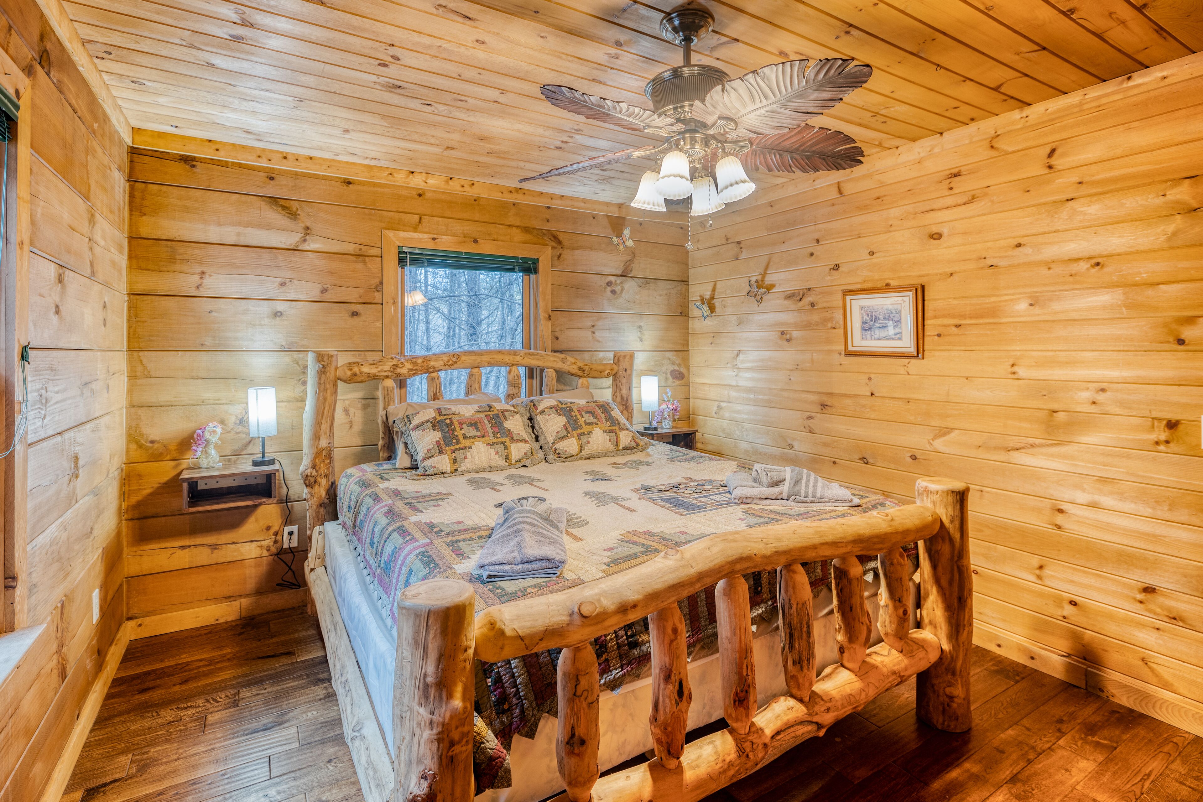 Une chambre dans une cabane en bois, avec un lit, une table de chevet, un ventilateur de plafond et une fenêtre.