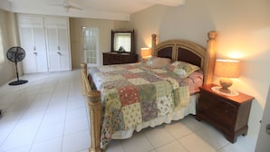 A bedroom with a bed, two bedside tables with lamps, a dresser, a ceiling fan, and a door with a fan beside it. - Spacious One Bedroom Apartment in Palmiste (San Fernando)