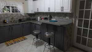 A kitchen with white cabinets, a granite countertop, two stools, a window with a curtain, and a stone backsplash. - Spacious One Bedroom Apartment in Palmiste (San Fernando)