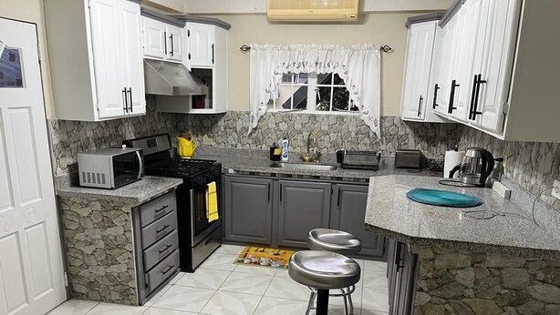 A kitchen with white cabinets, a stone backsplash, a gas stove, a microwave, a sink, and two bar stools. - Spacious One Bedroom Apartment in Palmiste (San Fernando)
