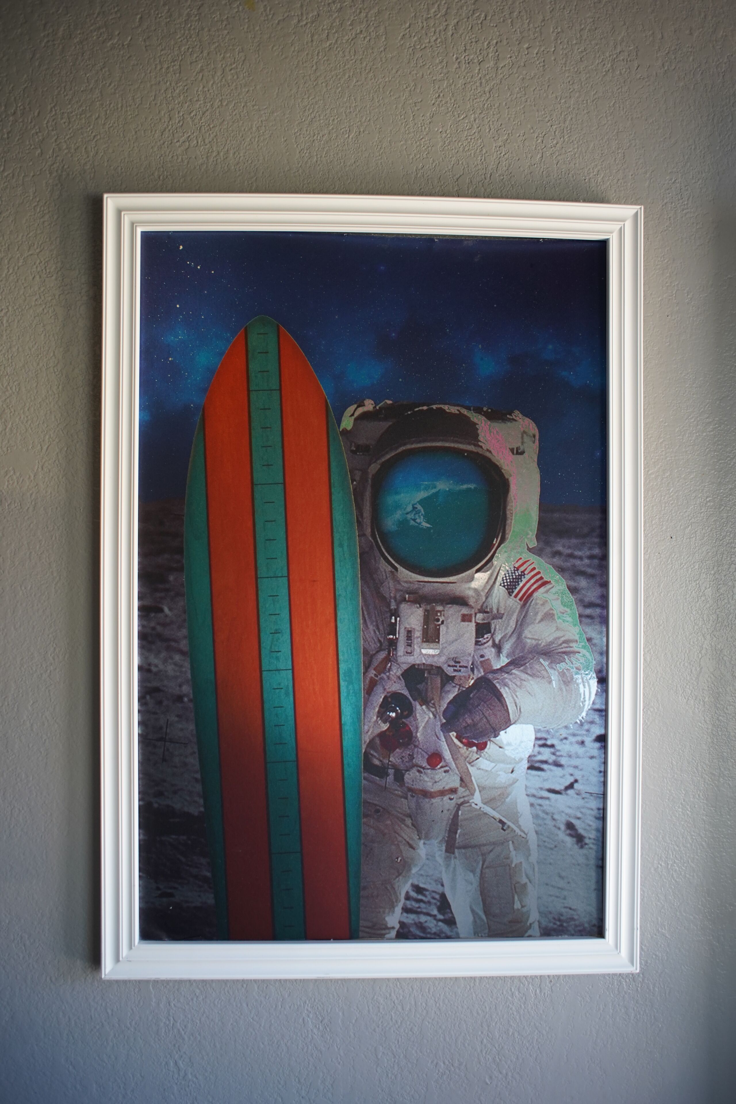A framed artwork of an astronaut holding a surfboard against a backdrop of space and Earth.