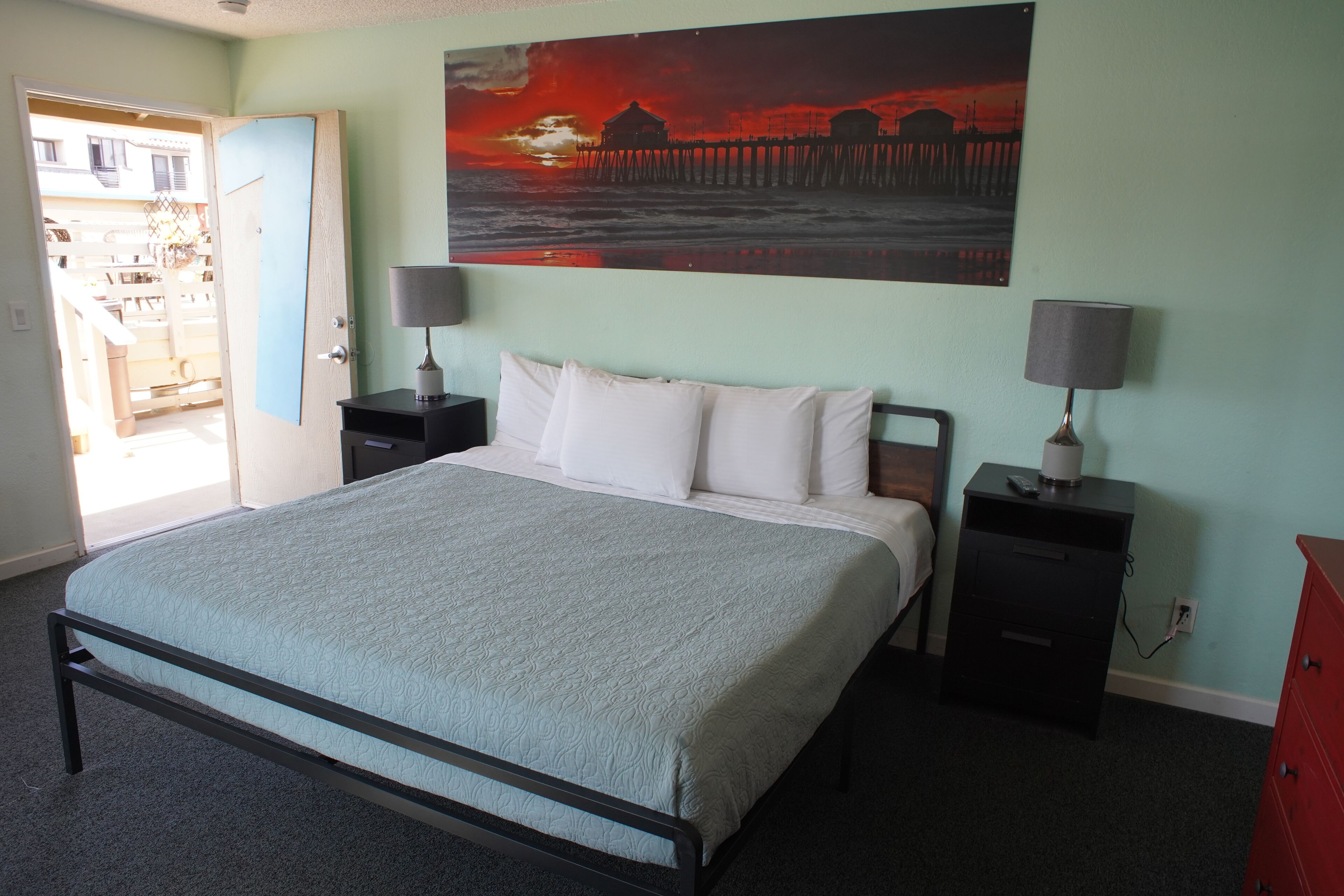A bedroom with a bed, two bedside tables, a nightstand, a red dresser, and a painting of a sunset over a pier.