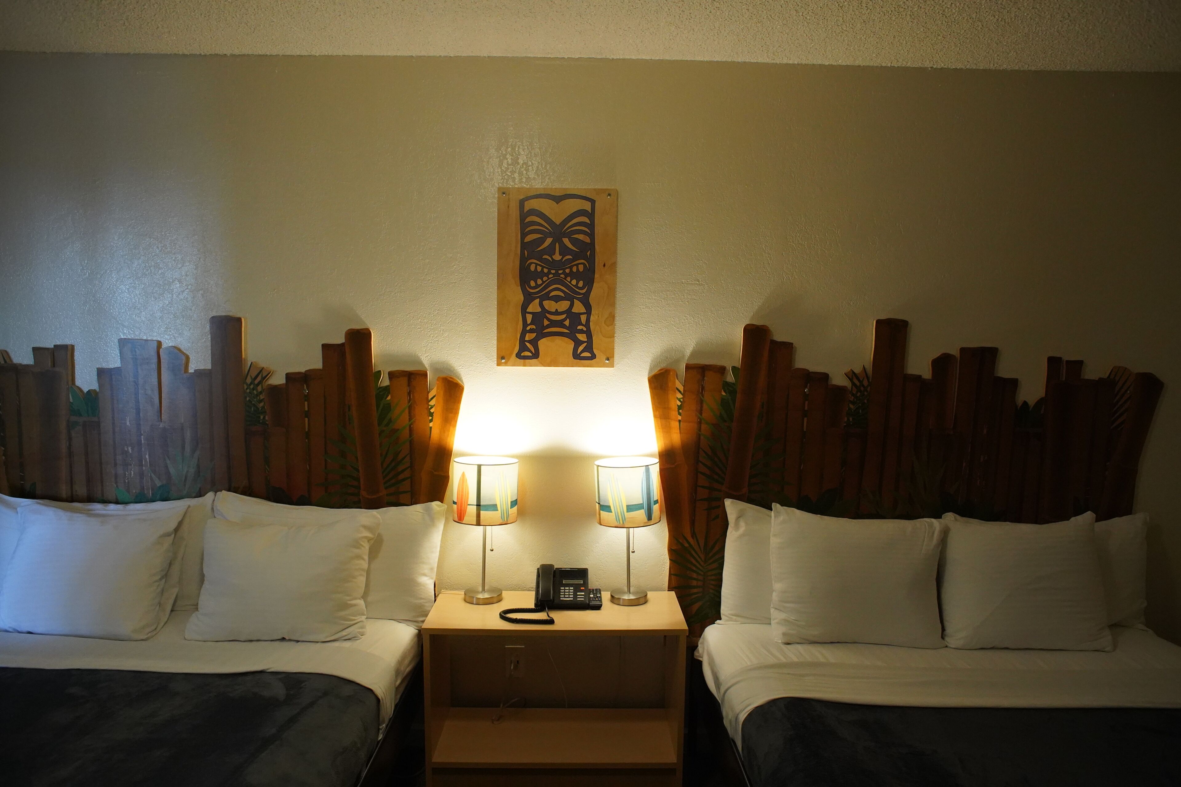 A hotel room with two beds, a nightstand with a phone, two lamps, and a wooden headboard with a tribal mask decoration.