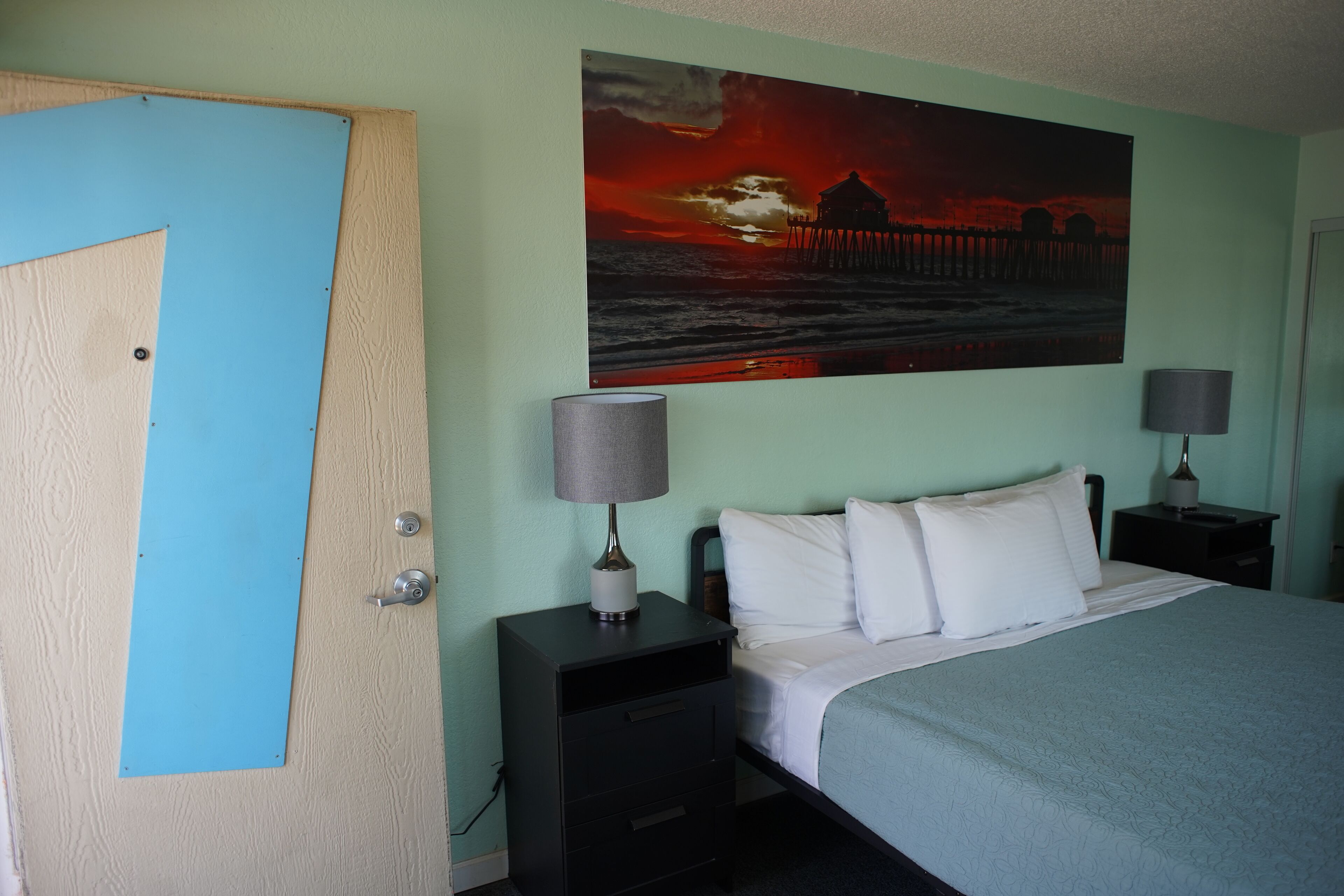 Un dormitorio con una cama, dos mesitas de noche, un cuadro de un atardecer y una puerta.