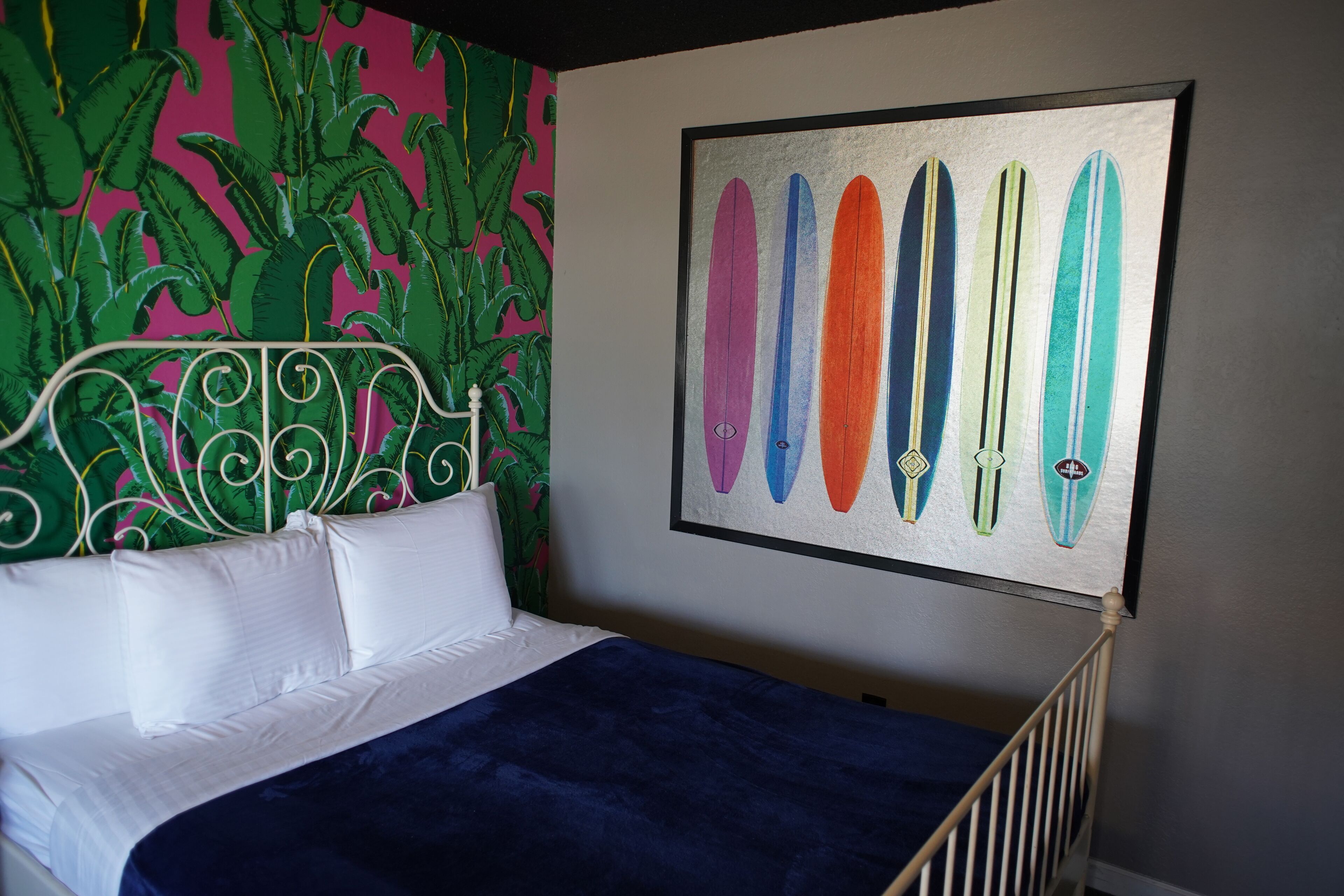 Ein Bett mit weißen Kissen und einer blauen Decke, ein Metall-Kopfteil und eine Wand mit einem gerahmten Surfbrett-Kunstwerk.