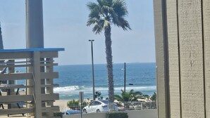 Une vue depuis un balcon donnant sur une plage avec un palmier, une rue bordée de voitures garées et un ciel dégagé.