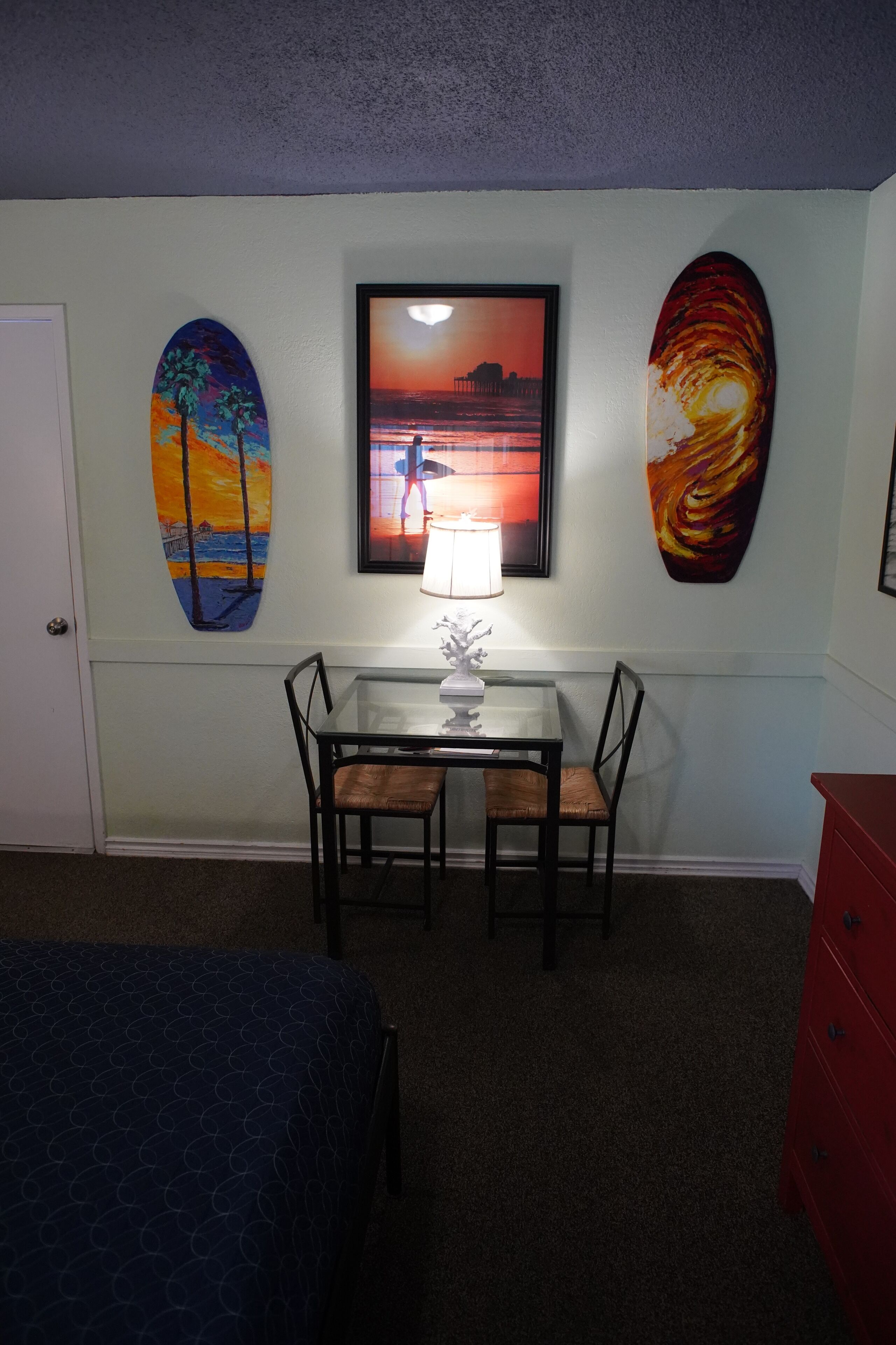 A room with a bed, a desk with two chairs, a lamp, and three paintings on the wall.