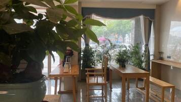 Una habitación con una planta grande en maceta, una mesa de madera, un banco y vistas a través de una ventana con cortinas.