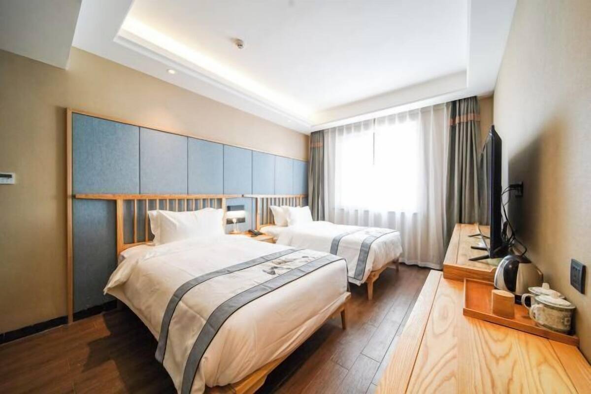 A hotel room with two beds, a wooden desk, a television, and a large window with curtains.