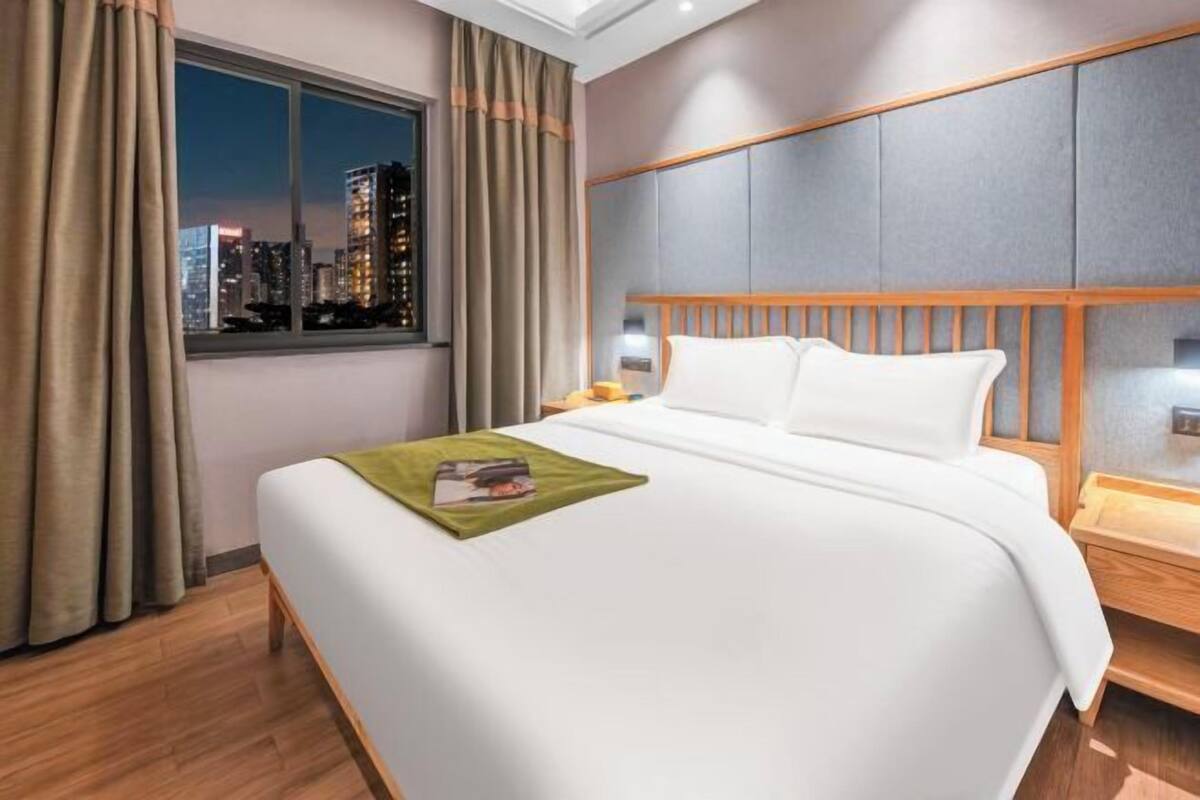 A hotel room with a large bed, a window with curtains, a nightstand, and a city view.
